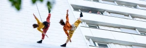 En contreplongée, deux acrobates font de la danse libre sur la façade d'un immeuble. Un voisin, depuis son balcon, regarde les circassiennes.