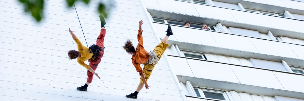En contreplongée, deux acrobates font de la danse libre sur la façade d'un immeuble. Un voisin, depuis son balcon, regarde les circassiennes.