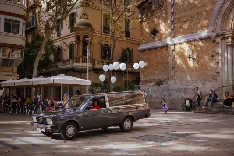 Un corbillard est stationné sur une place ; huit ballons blancs sont suspendus sur sa carrosserie. Une femme vêtue de rouge est au volant.