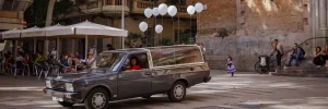 Un corbillard est stationné sur une place ; huit ballons blancs sont suspendus sur sa carrosserie. Une femme vêtue de rouge est au volant.