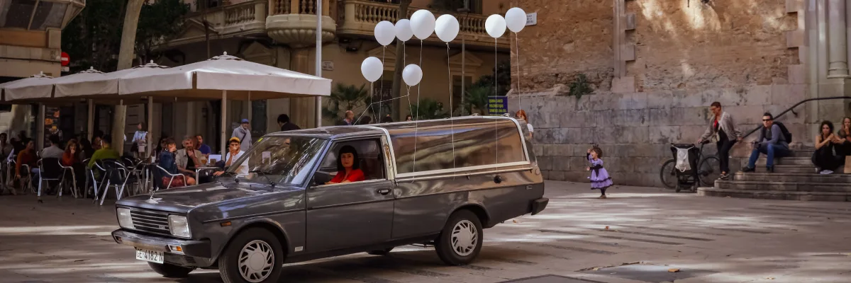 Un corbillard est stationné sur une place ; huit ballons blancs sont suspendus sur sa carrosserie. Une femme vêtue de rouge est au volant.